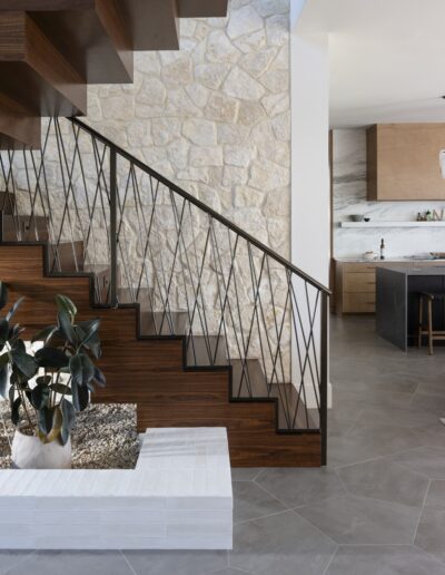 Modern interior with a wooden staircase featuring black metal railings, stone accent wall, gray tile floor, and a small indoor planter with a green plant. Kitchen visible in the background.