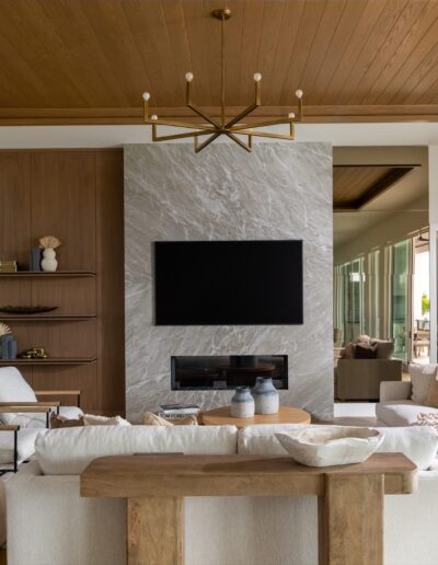 Modern living room with a large wall-mounted TV, fireplace, light furniture, wooden accents, and large windows letting in natural light.
