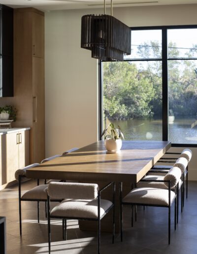 A modern dining room with a wooden table, six upholstered chairs, a black pendant light, and a large window overlooking water and trees.
