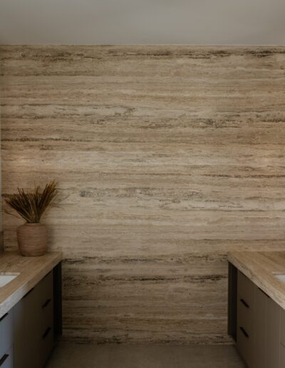 Modern bathroom with double sinks, travertine countertops and backsplash, gold fixtures, large mirrors, and wall-mounted lights on each side. A vase with dried plants and a tray with towels are on the counters.