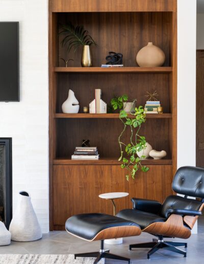 A modern living room features a wood bookshelf with decor items, a black lounge chair with ottoman, a wall-mounted TV, and a fireplace.