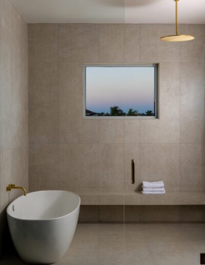 Minimalist bathroom with a white freestanding tub, gold fixtures, folded white towels on a bench, beige tile walls, and a small window showing an evening sky.