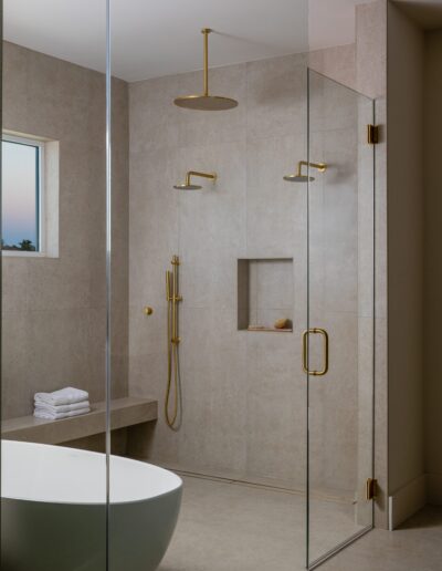 Modern bathroom with a freestanding bathtub, a glass-enclosed shower featuring gold fixtures, a built-in shower niche, beige tile walls, a bench, and a window.