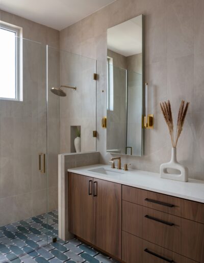 Modern bathroom with wood vanity, rectangular white sink, gold fixtures, glass shower enclosure, and blue tiled floor. A vase with dried pampas grass sits on the counter.