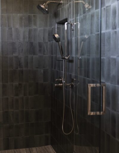 Modern shower with dark rectangular tiles, a glass door, a wall-mounted showerhead, a handheld shower, and metal fixtures.