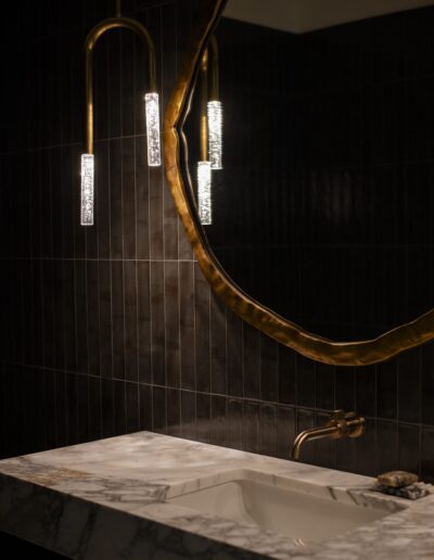 A modern bathroom features a marble sink, a gold faucet, a large round mirror with an irregular gold frame, and pendant lights against dark tiled walls.