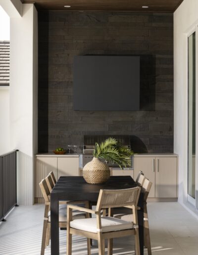 A modern patio with a black dining table, four wooden chairs, a woven vase with green leaves, built-in cabinetry, a TV on a dark accent wall, and a ceiling fan overhead.