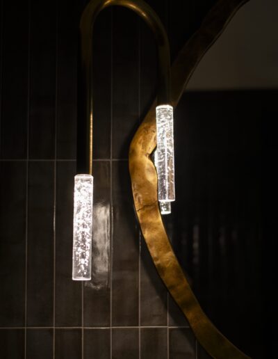 A modern hanging light fixture with two cylindrical illuminated bulbs is next to a curved, gold-edged mirror against a dark tiled wall.