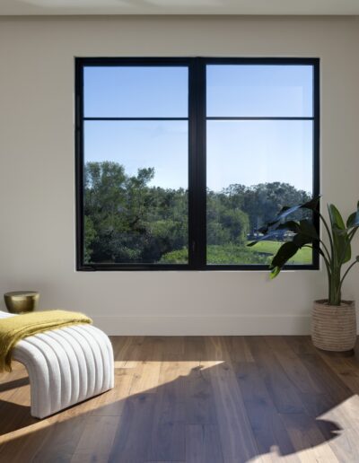 A modern, minimalist room with a sculptural white chaise lounge, a yellow throw, a potted plant, wood floors, and large windows overlooking greenery.