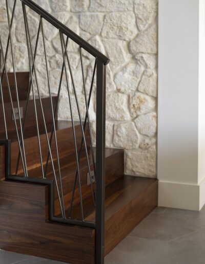 Dark wood staircase with black metal railing set against a light-colored stone wall and gray tile flooring.