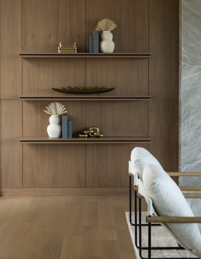 Three wooden floating shelves with decorative vases, books, a sculptural object, and a bowl are mounted on a wood-paneled wall beside two upholstered chairs.