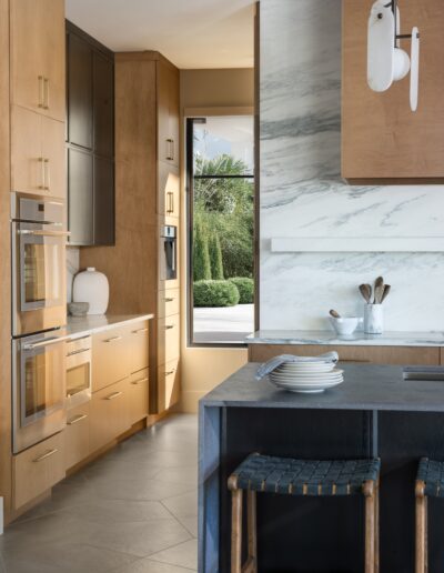 Modern kitchen with light wood cabinets, marble backsplash, built-in ovens, and a dark island with stools; large window shows greenery outside.
