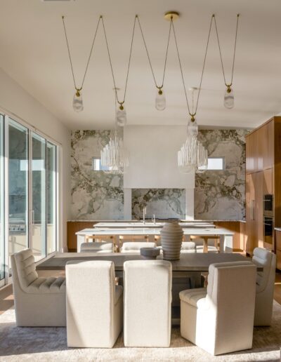 Modern dining room with beige upholstered chairs, a wooden table, pendant lights, large windows, marble accents, and built-in wooden cabinetry.