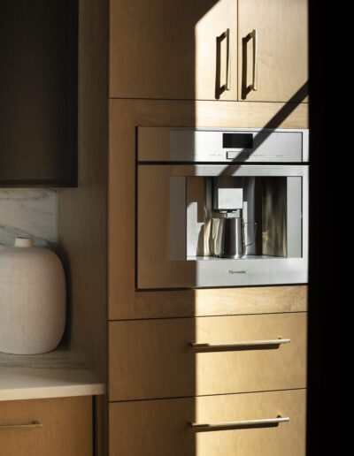 Built-in stainless steel coffee machine set into light wood cabinetry, with a silver carafe under the dispenser and sunlight casting a shadow across the scene.