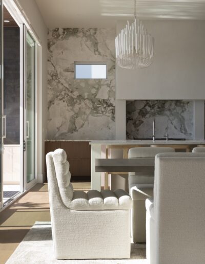 Modern dining area with tufted cream chairs, a rectangular table, marble accent wall, large sliding glass doors, and a white sculptural chandelier. Natural light fills the space.
