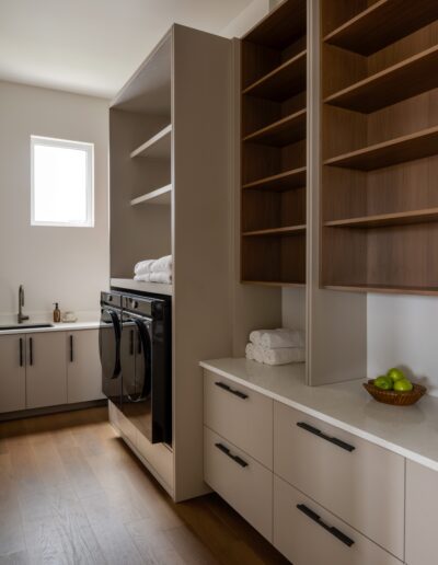 Modern laundry room with light wood flooring, beige cabinetry, open shelving, stacked black washer and dryer, small sink, folded towels, and a bowl of green apples on the counter.
