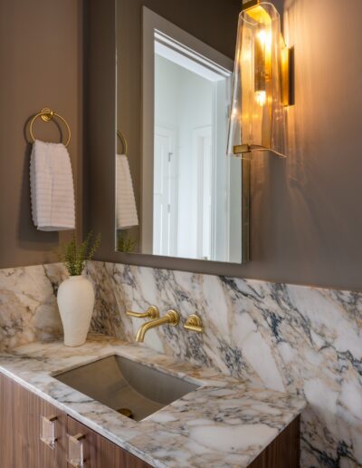 Modern bathroom with a marble countertop, built-in sink, wall-mounted faucet, gold accents, a white vase, and a lit glass wall sconce beside a mirror.