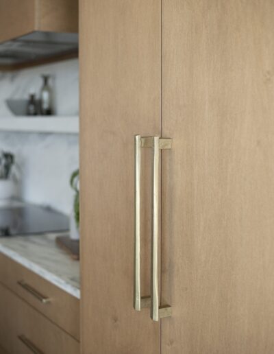 Close-up of two gold handles on a light wood cabinet door in a modern kitchen with a marble countertop and open shelving in the background.
