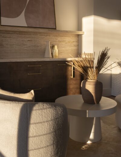 A sunlit modern living room features a round white coffee table with a ceramic vase of dried plants, a beige armchair, and dark wood cabinets against a textured wall.