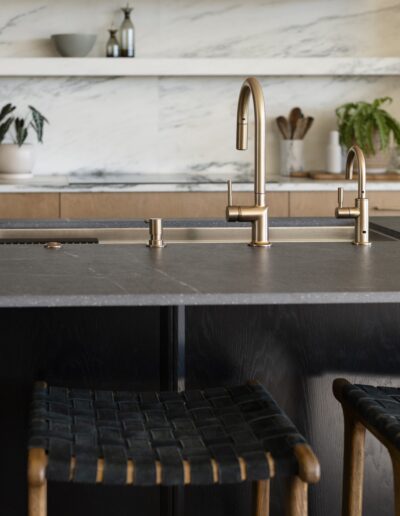Modern kitchen with a marble backsplash, open shelves, gold faucets on a dark island countertop, and two woven barstools in the foreground.