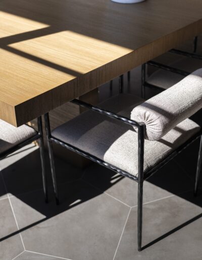 A close-up of a wooden table with metal-framed chairs featuring padded seat and back cushions, on a gray tiled floor with sunlight casting shadows.