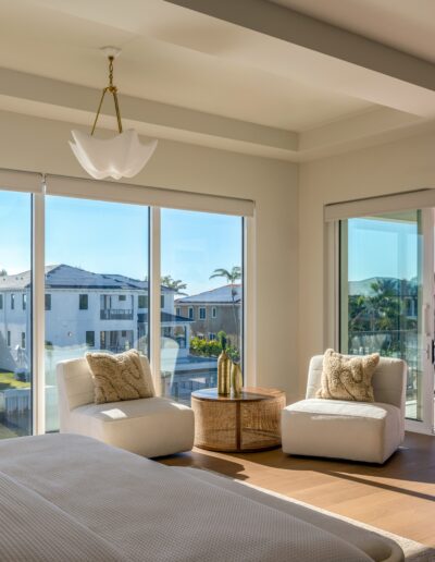 A modern bedroom with large windows, two chairs, a small table, and a balcony overlooking neighboring houses in a sunny residential area.